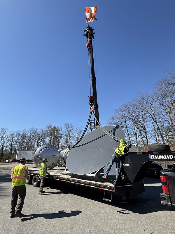 Workers in safety vests unload a large, silver, cylindrical piece of equipment from a flatbed truck, with a crane lifting another similar object, against a clear blue sky and trees in the background.