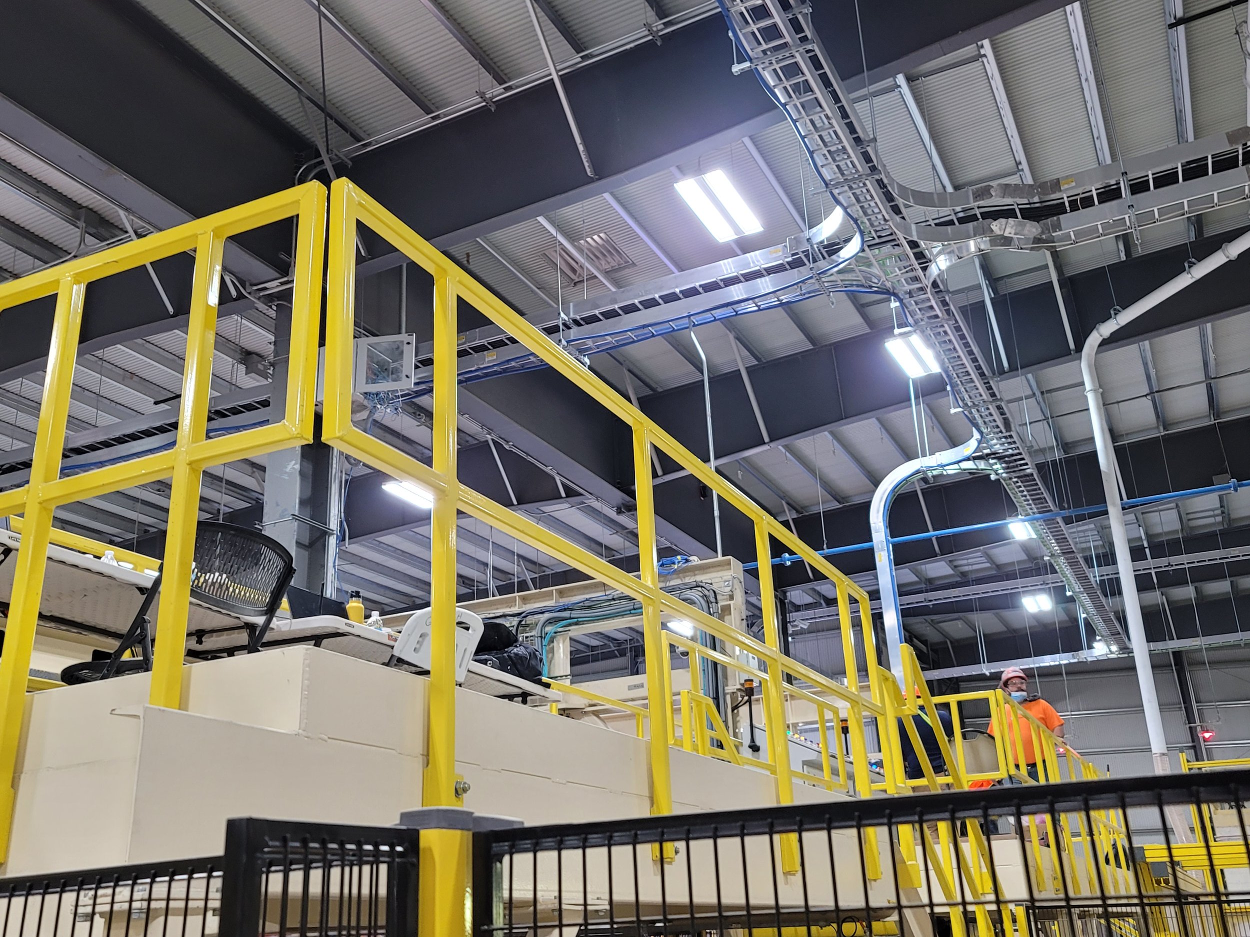 Interior of a warehouse or industrial facility with yellow safety railings, overhead track lighting, and various electrical and cable conduits running along the ceiling. There are people working in the background, one wearing an orange shirt and a fa