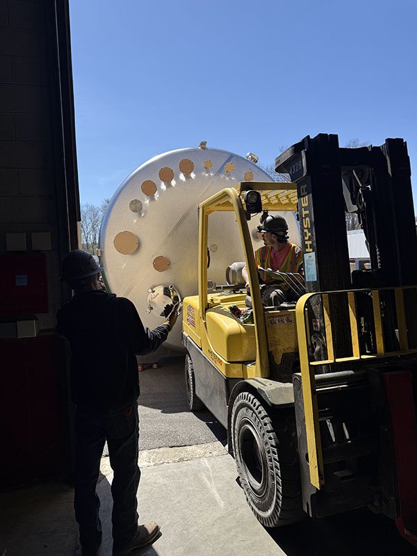 Workers using a forklift to move a large, cylindrical industrial tank outside.