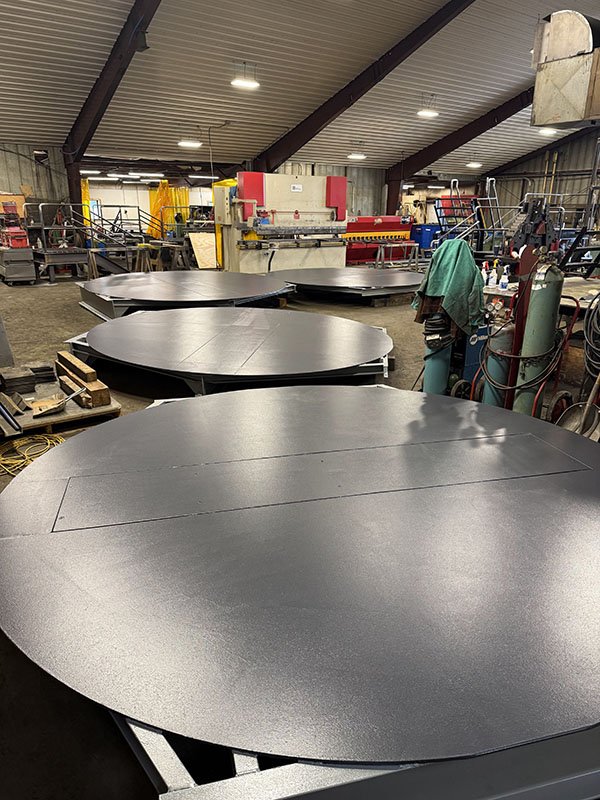 Large black circular metal sheets on worktables in a factory or workshop with industrial equipment and tools in the background.