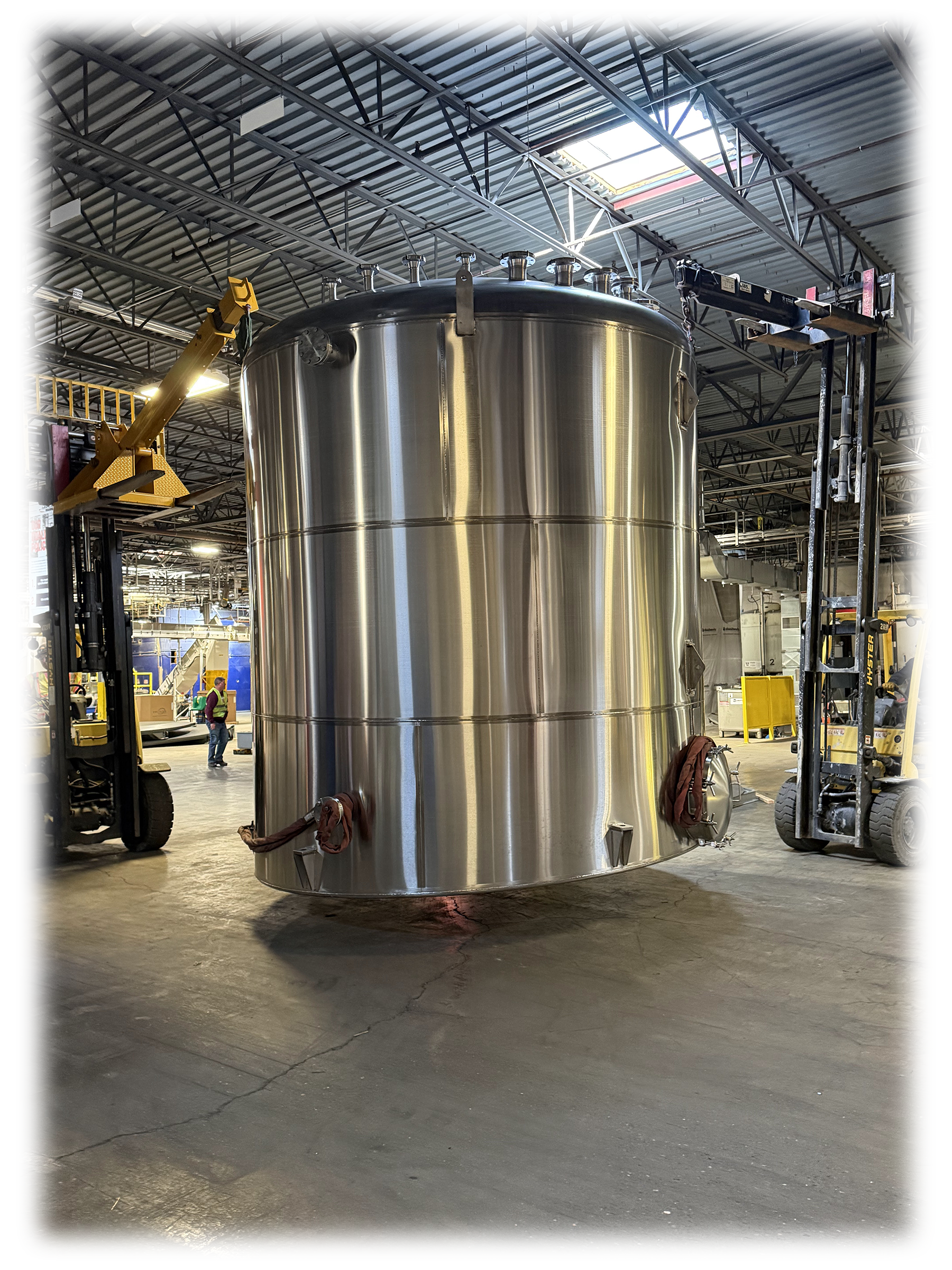 Large stainless steel cylindrical tank suspended in an industrial warehouse, with lifting equipment and workers in the background.