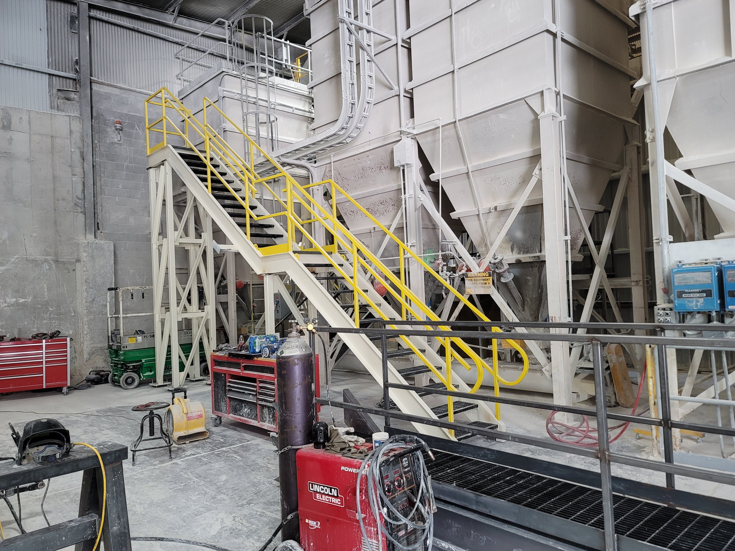 Industrial workshop with metal stairs featuring yellow safety railings, large white machinery, tools, and equipment scattered around, including a red Lincoln Electric welder, and a warning sign visible among supplies.
