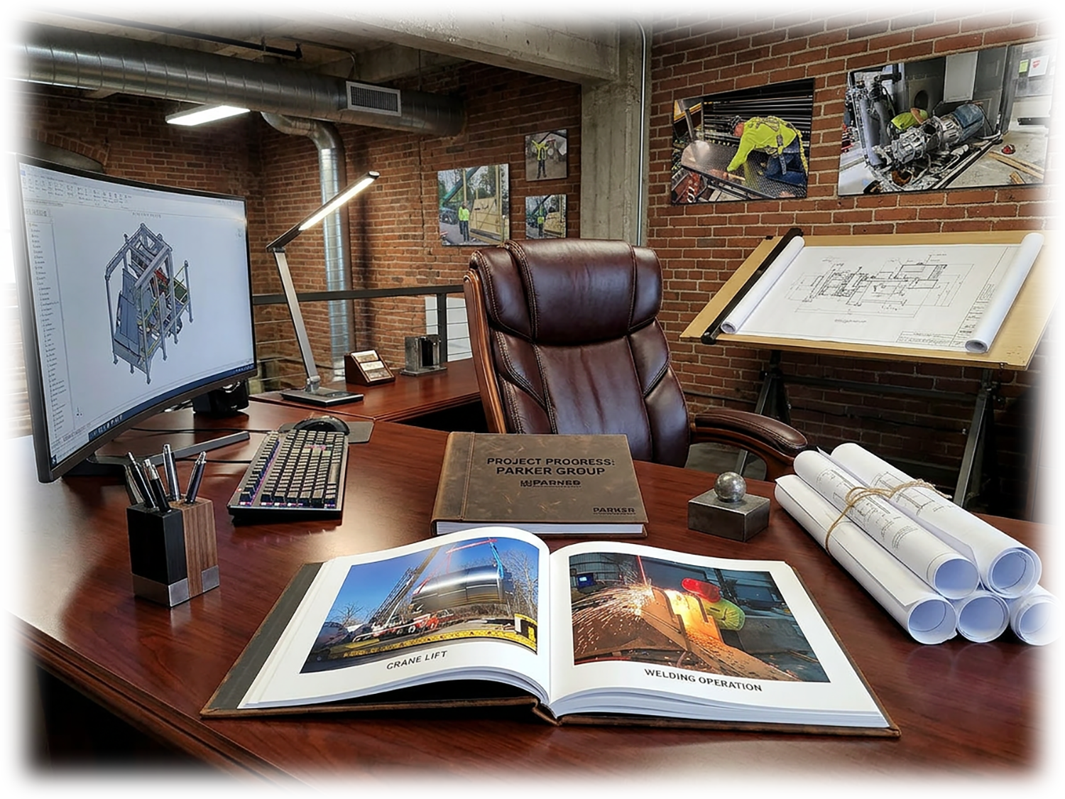 Office desk with a computer, architectural blueprints, construction project manuals, and images of construction activities on the wall and monitor.
