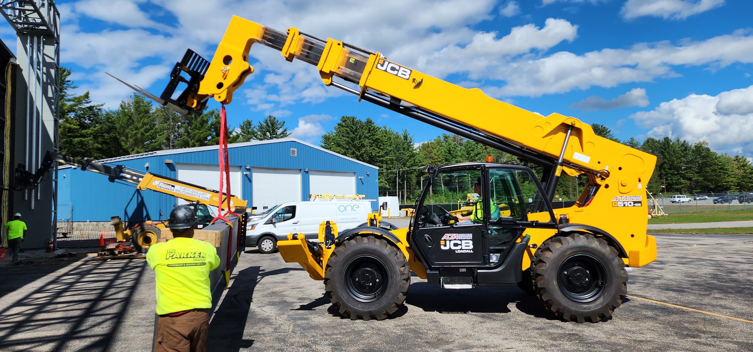 A yellow JCB telescopic boom lift lifting a large red strap, with workers and vehicles nearby at an outdoor construction or loading site.