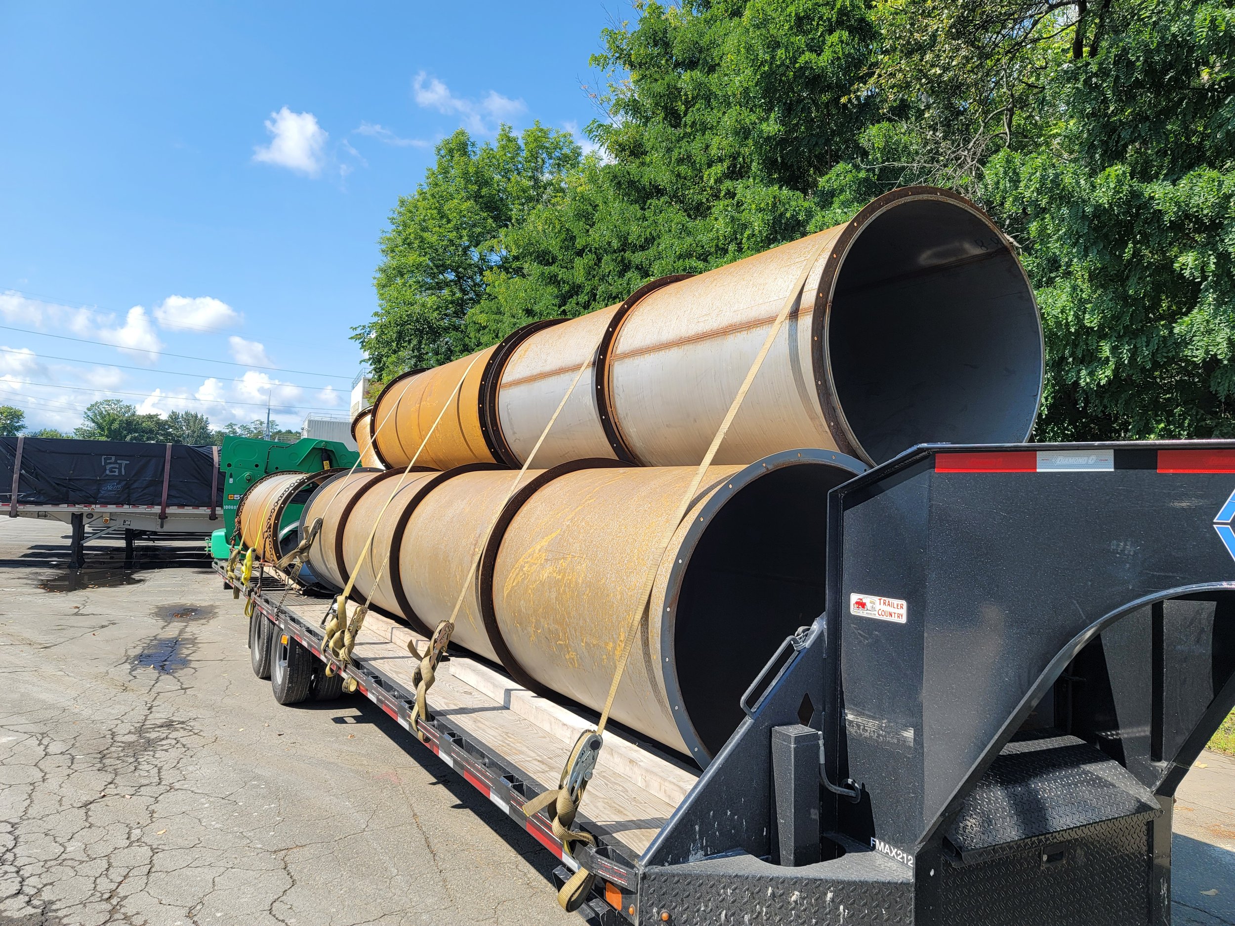 A flatbed trailer loaded with large metal pipes with a rusty exterior, parked on a cracked asphalt surface next to green trees under a blue sky with some clouds.