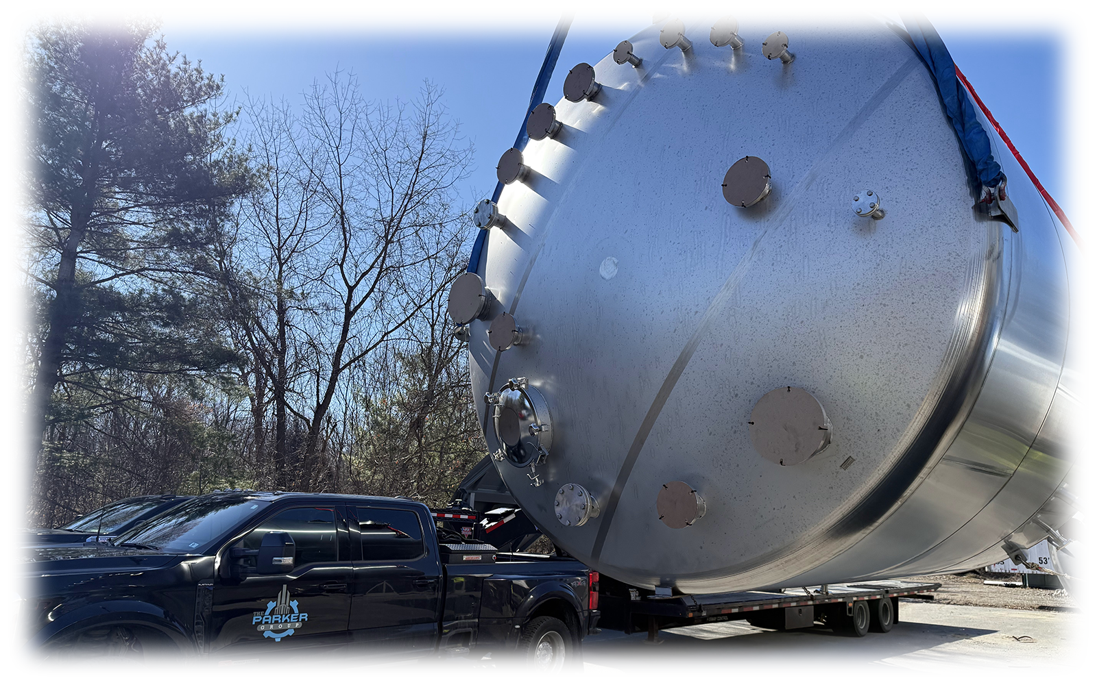 A large cylindrical tank is loaded onto a flatbed truck. The tank appears to be metallic and reflective, with several circular access ports or hatches.