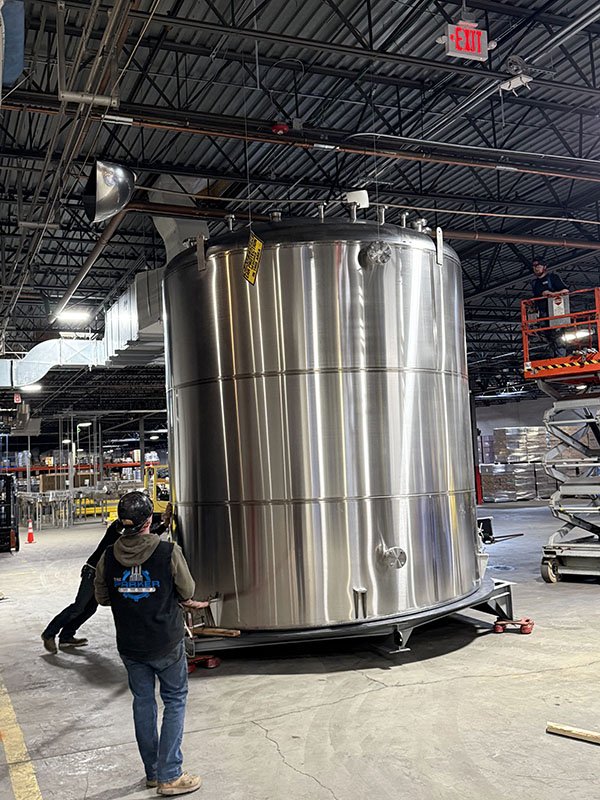 Large stainless steel industrial storage tank inside a warehouse, with workers and equipment around it.