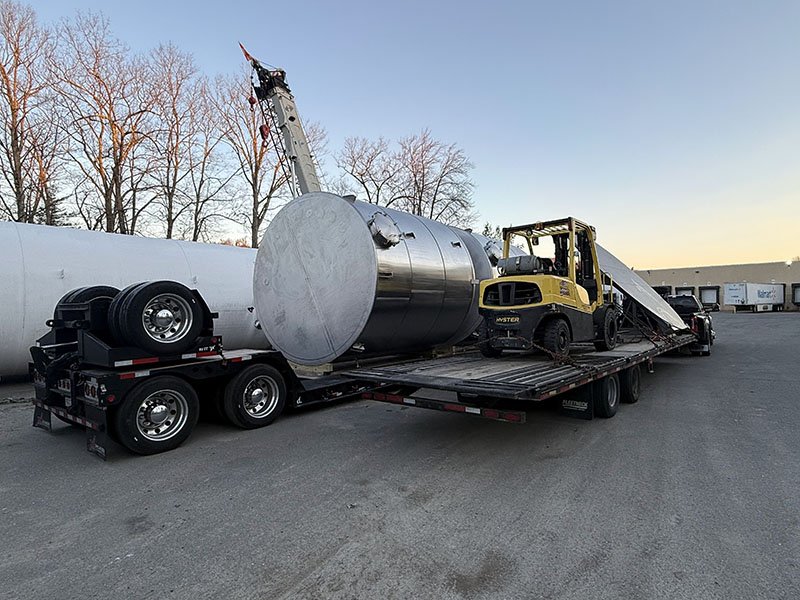 A flatbed truck transporting a large stainless steel tank, a yellow forklift, and a long white industrial pipe in an industrial area with a clear sky and leafless trees in the background.