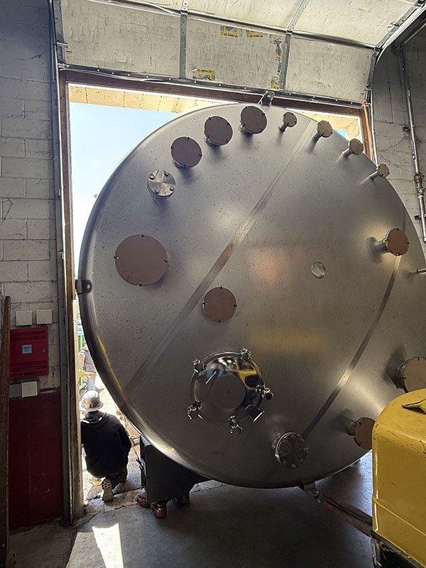 Large stainless steel industrial tank being moved into a warehouse or factory through a wide open door. The tank has several circular access ports and fitting attachments on its surface.