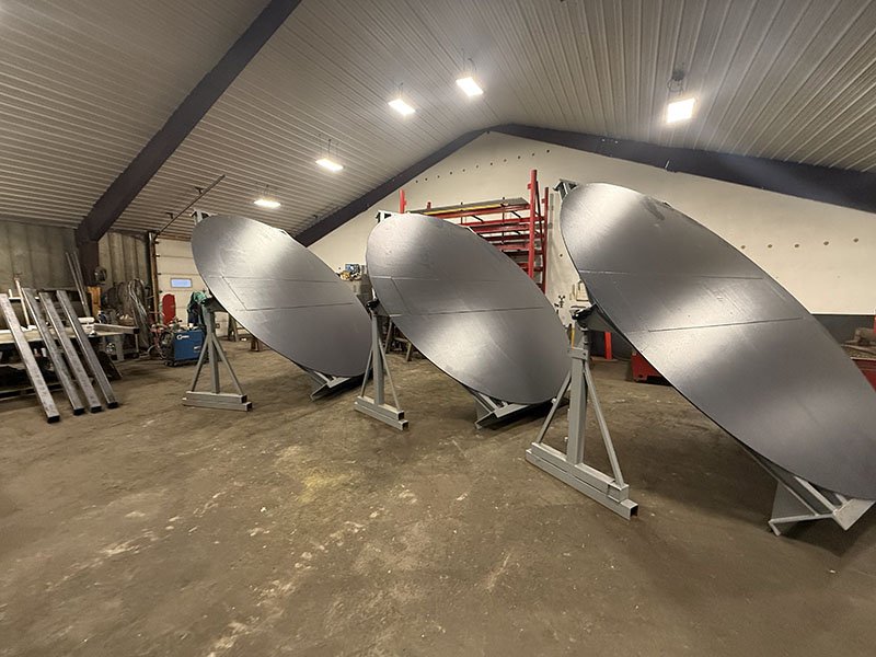 Three large satellite dishes inside a warehouse or workshop, mounted on metal frames, with tools and equipment in the background.
