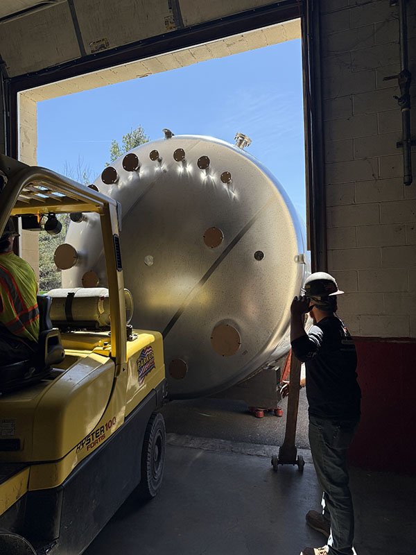 A large cylindrical object with multiple circular openings being moved by a yellow forklift inside a building with a large open door, a person standing nearby wearing a hard hat and glasses.
