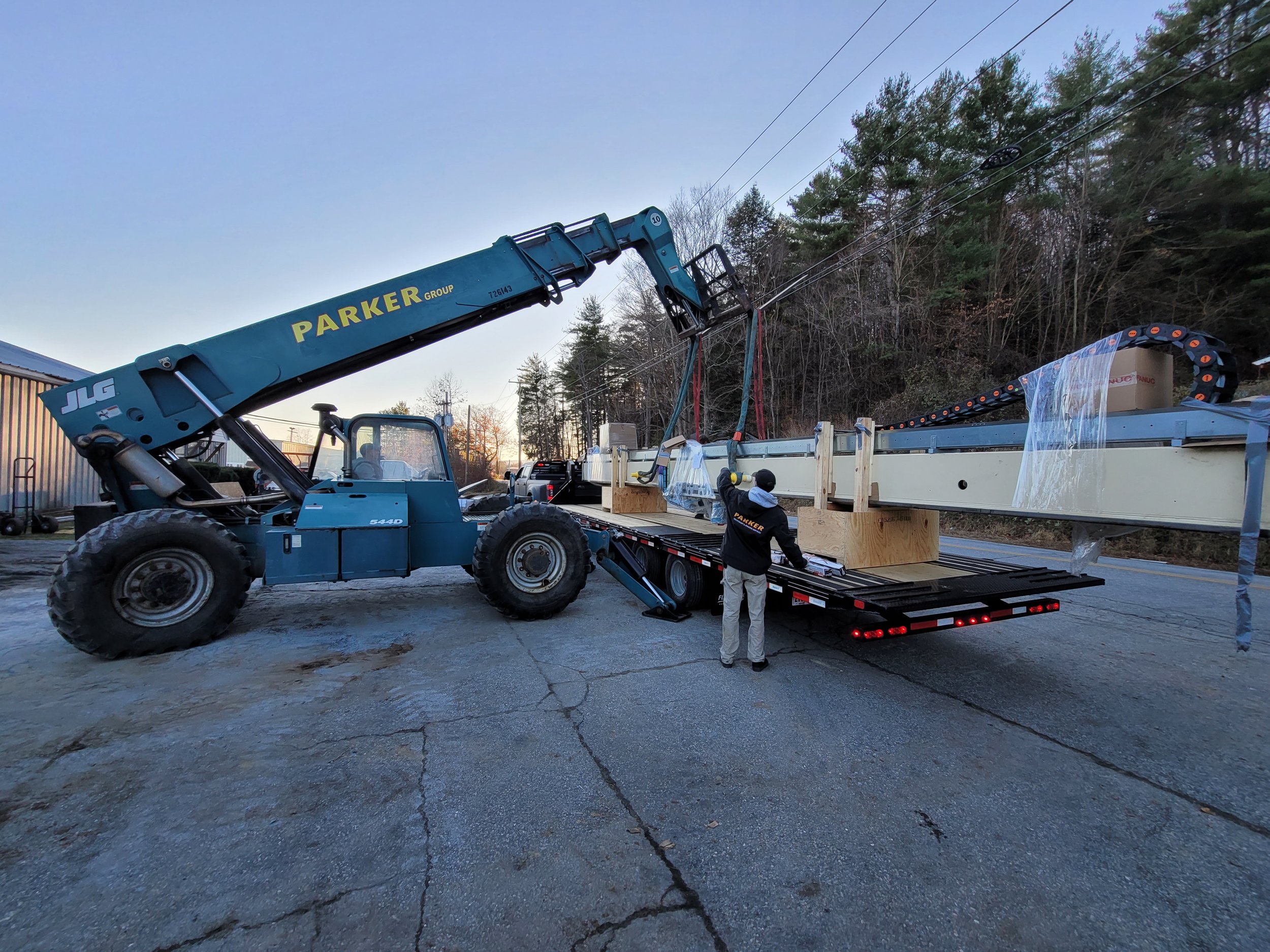 A large blue crane lifting a long metal beam onto a flatbed trailer, with two workers guiding the process outdoors near trees and power lines at sunset.