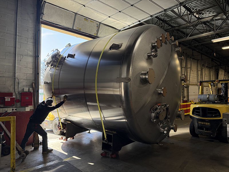 A worker is standing near a large cylindrical metal tank inside a warehouse, with a yellow forklift nearby.