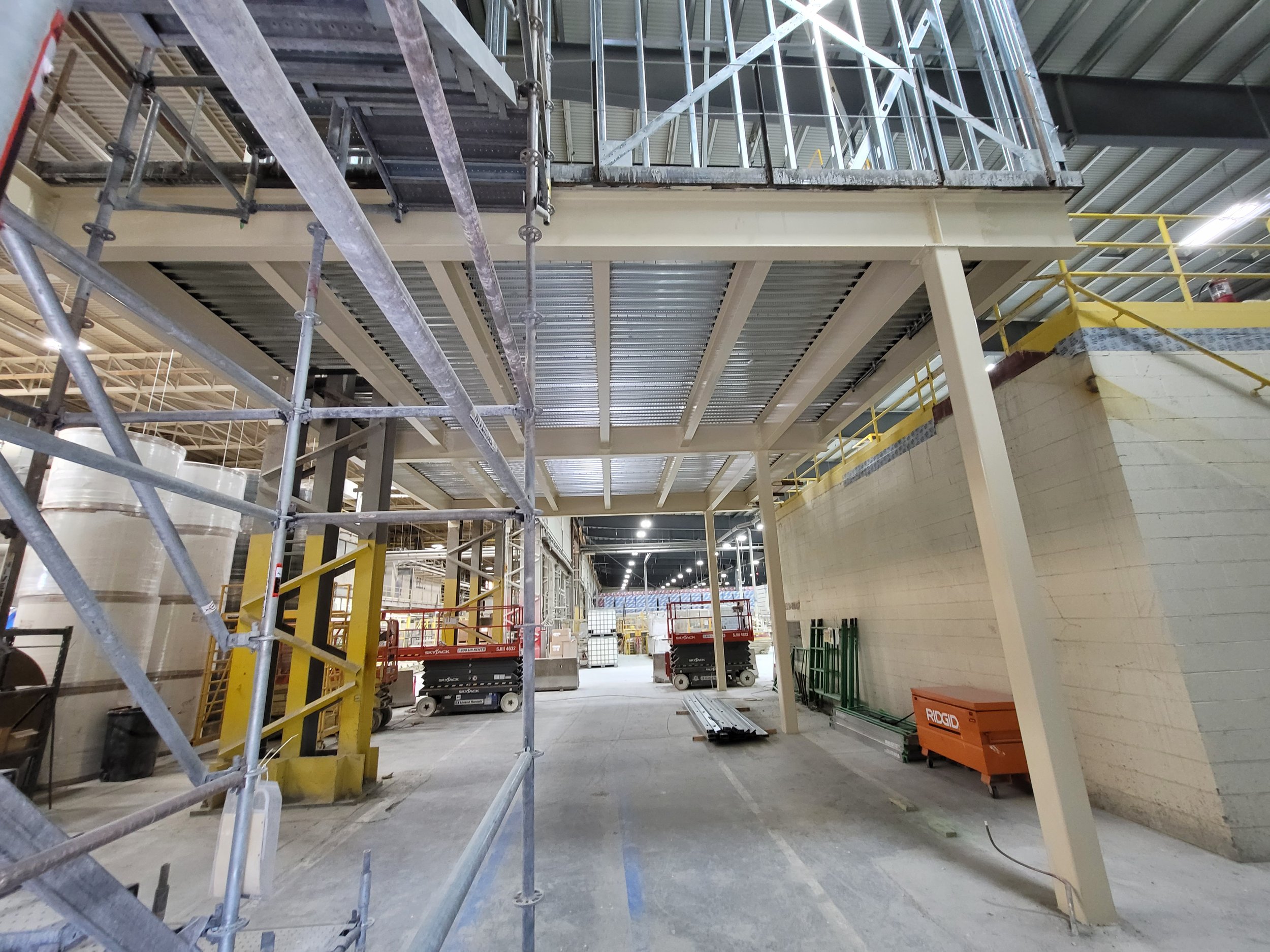 Construction inside a building with scaffolding, yellow safety rails, and construction equipment, including scissor lifts and a work platform.