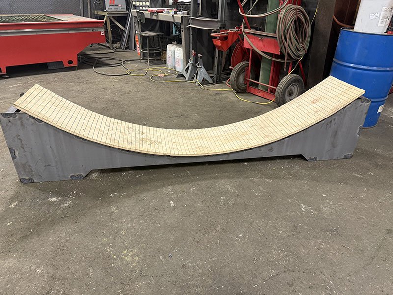 A wooden skateboard ramp with a curved design resting on a flat, gray base in a workshop or garage with tools and supplies in the background.