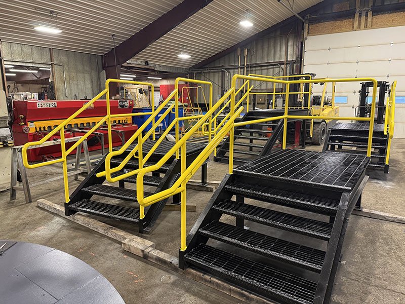Industrial warehouse with staircases and ramps, yellow safety railings, metal flooring, and equipment, including a yellow vehicle in the background.