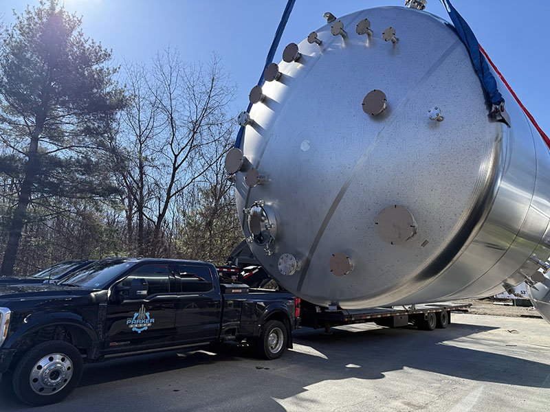 A large industrial stainless steel tank on a flatbed trailer being transported by a black pickup truck.