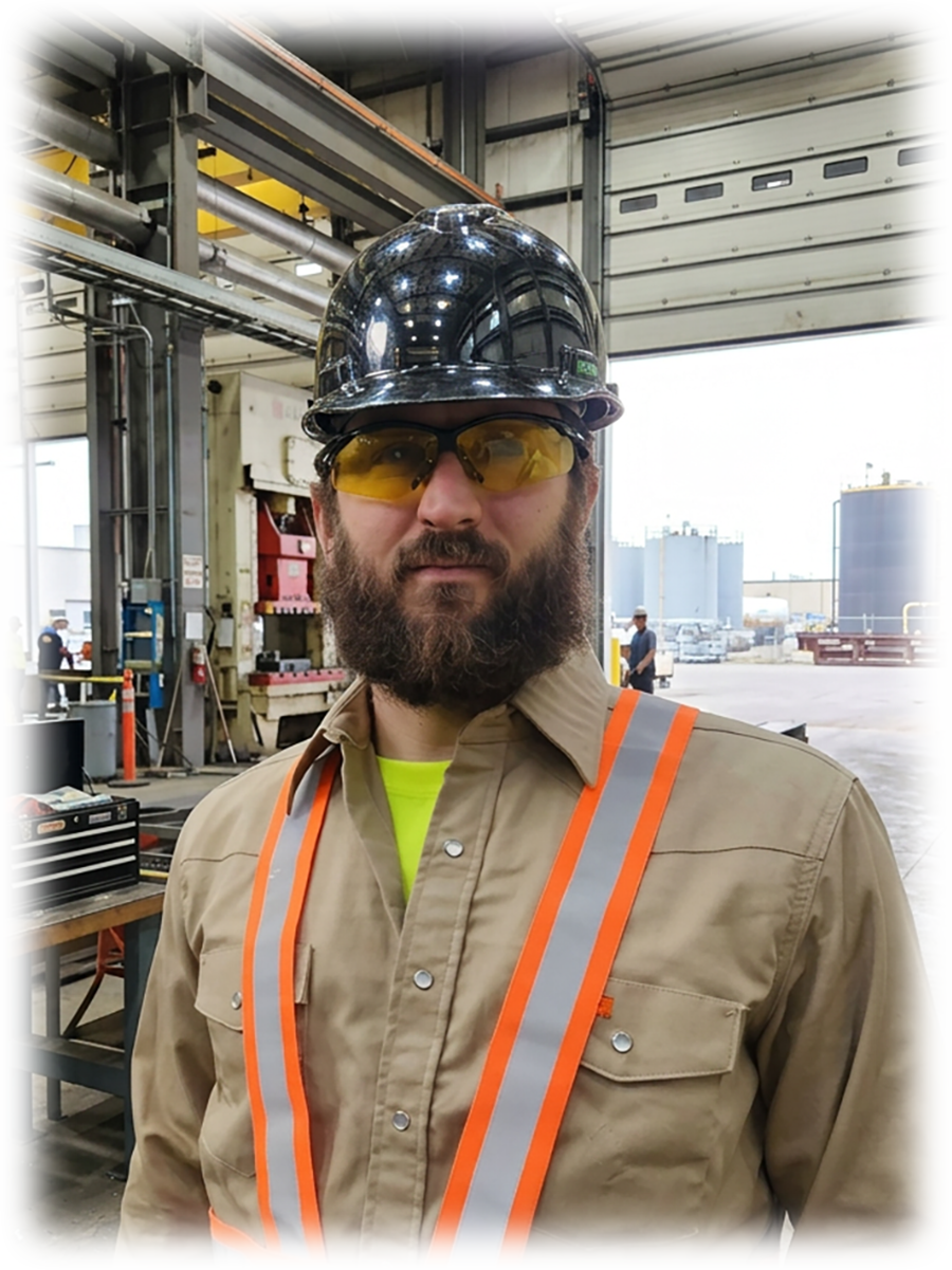 A man wearing a black hard hat, yellow safety glasses, and a tan work shirt with reflective orange and silver safety vest inside an industrial warehouse or manufacturing facility.