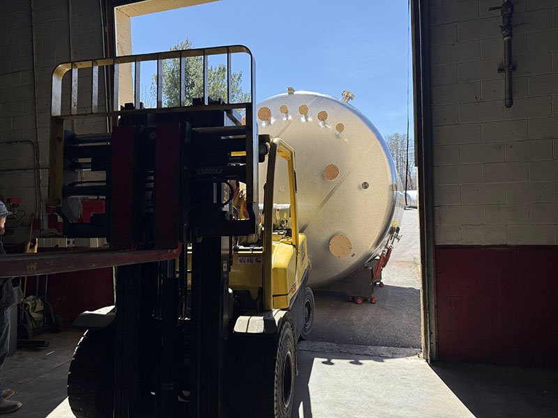 A forklift inside a warehouse next to a large horizontal storage tank outside.