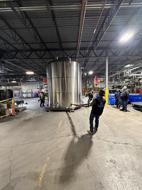 Industrial warehouse with a large stainless steel tank in the center, several workers around, some near the tank, others in the background, and various equipment and boxes on the sides.