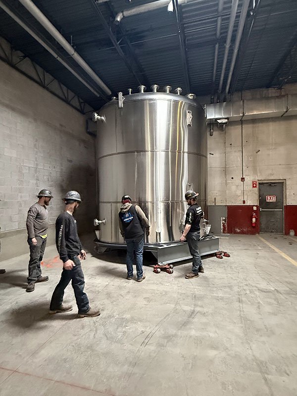 Five workers wearing helmets and work clothes observe a large stainless steel industrial tank in an indoor facility with concrete walls and a blue ceiling.