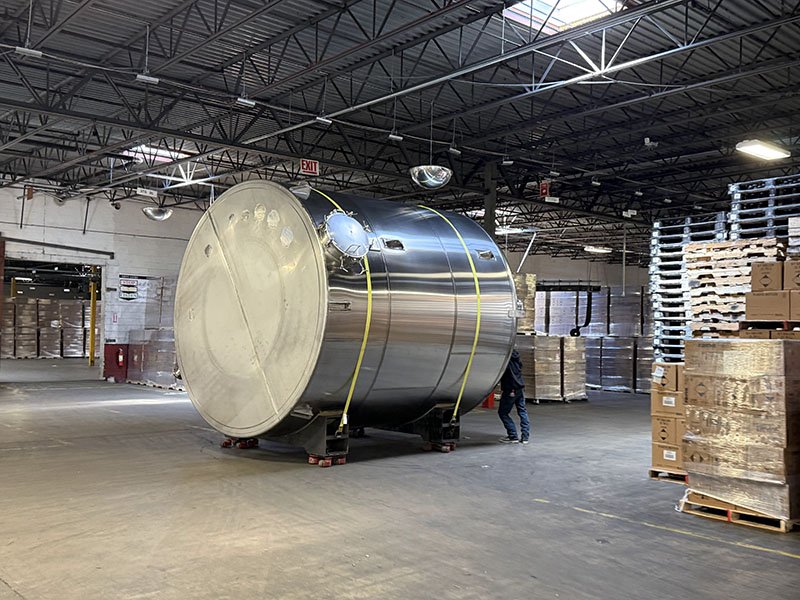 Large stainless steel industrial tank in a warehouse surrounded by stacked pallets and boxes.