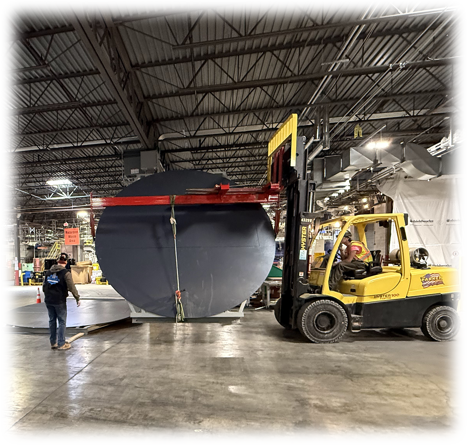 Workers use a forklift to move large cylindrical objects inside a warehouse. The warehouse has a high ceiling with exposed beams and ventilation ducts.