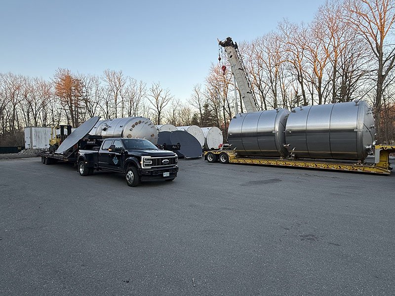 A large industrial crane lifting a massive, cylindrical stainless steel tank onto a flatbed trailer, with two additional tanks nearby, in an outdoor area with leafless trees and a clear sky.