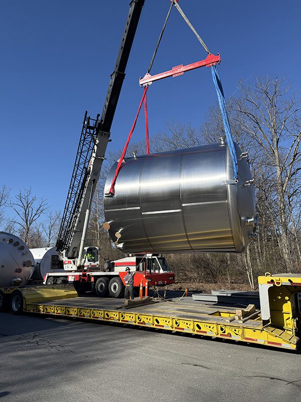 A large cylindrical stainless steel tank being lifted by a crane onto a flatbed truck. The scene takes place outdoors on a clear, sunny day with leafless trees in the background.