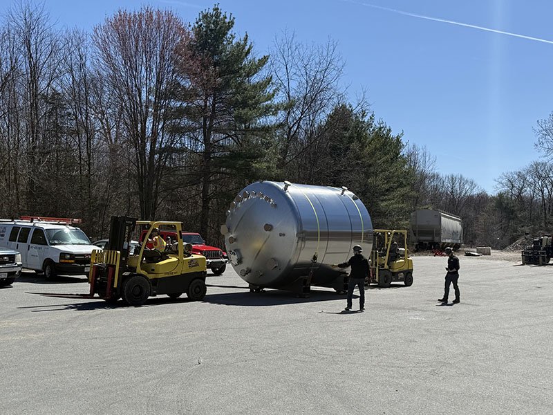 Three people and two forklifts move a large metallic cylindrical tank in a parking lot, with parked cars and trees in the background.
