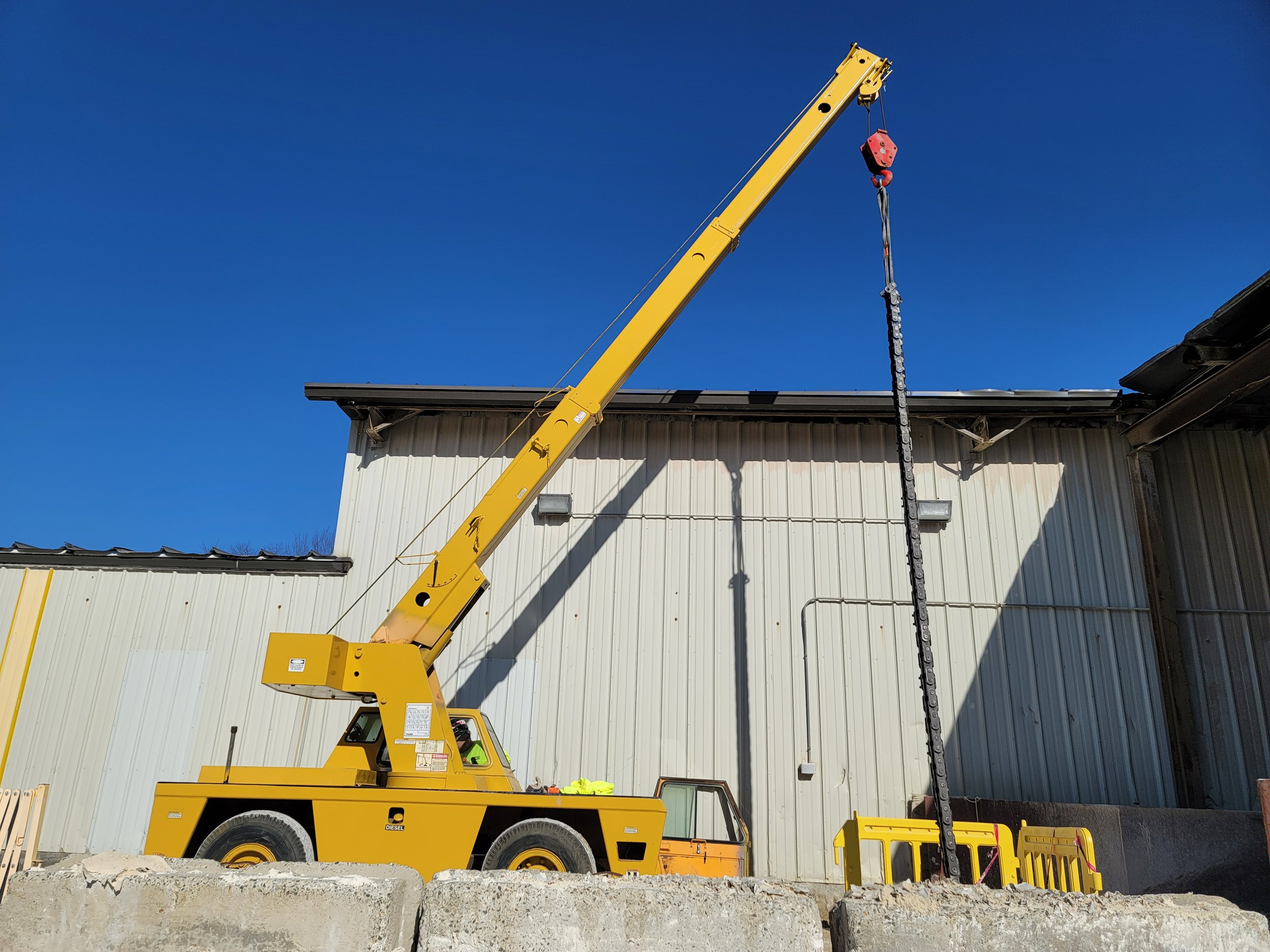 A yellow crane truck with a long extension arm elevates a chain hoist hanging from a building roof edge. The truck is parked on an uneven dirt surface. The background features a white metal building with a black roof, under a clear blue sky.