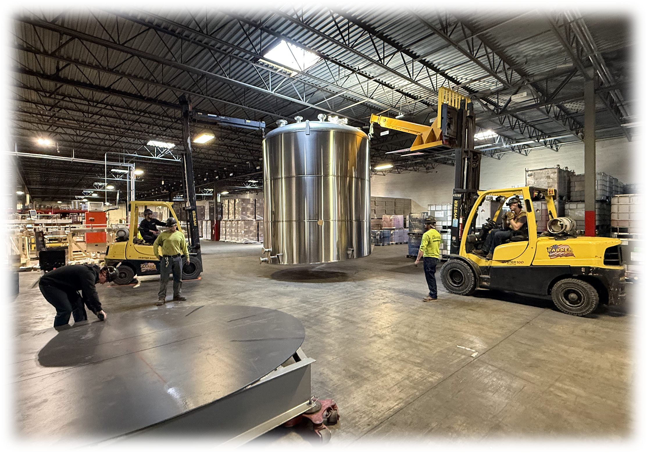Workers operate forklifts in an industrial warehouse to move large, shiny stainless steel tanks or containers.