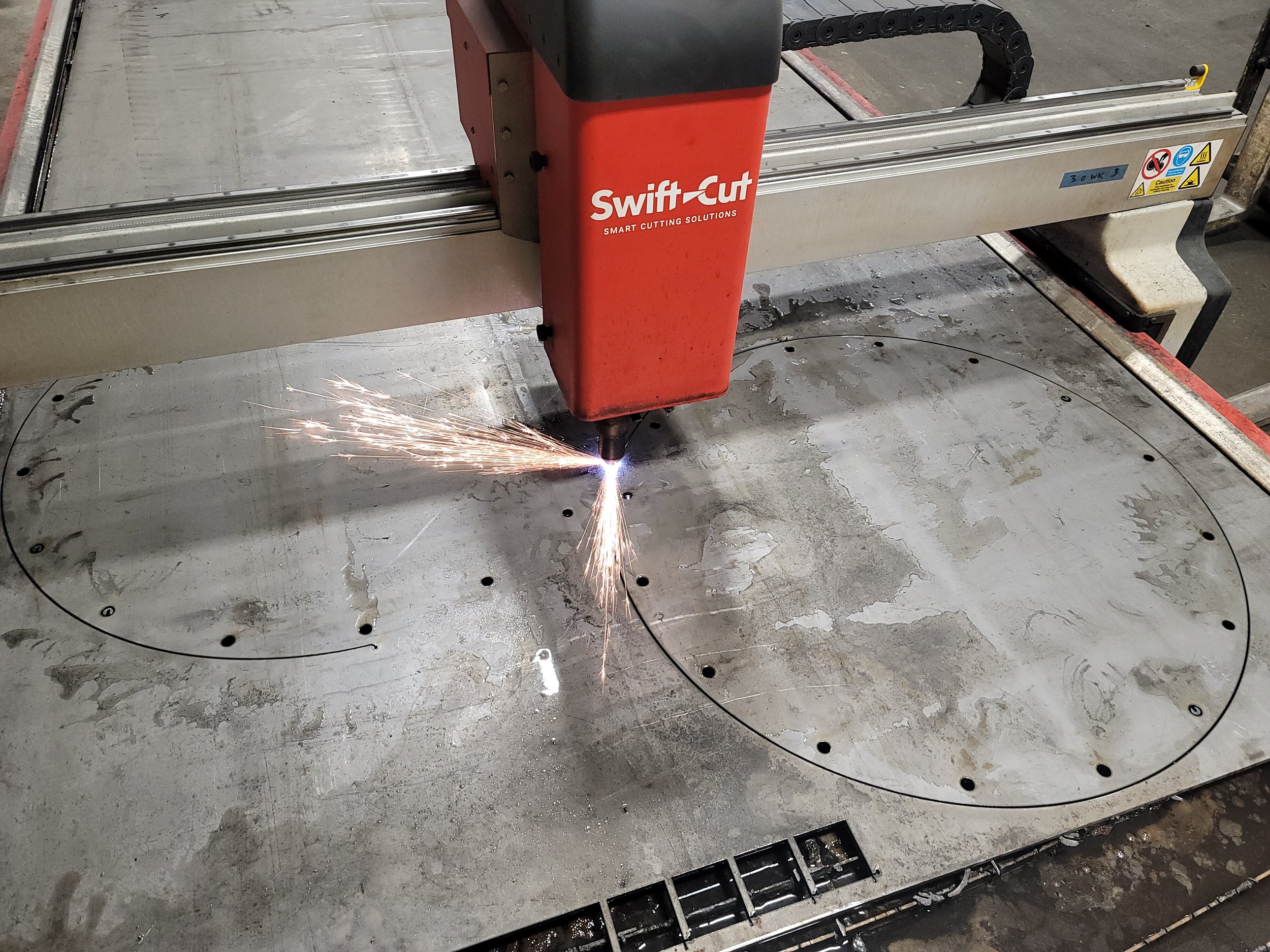 A CNC plasma cutting machine performing a cut on a metal circular plate, with sparks flying from the cut. The machine is labeled 'SwiftCut' and is surrounded by warning and safety stickers.