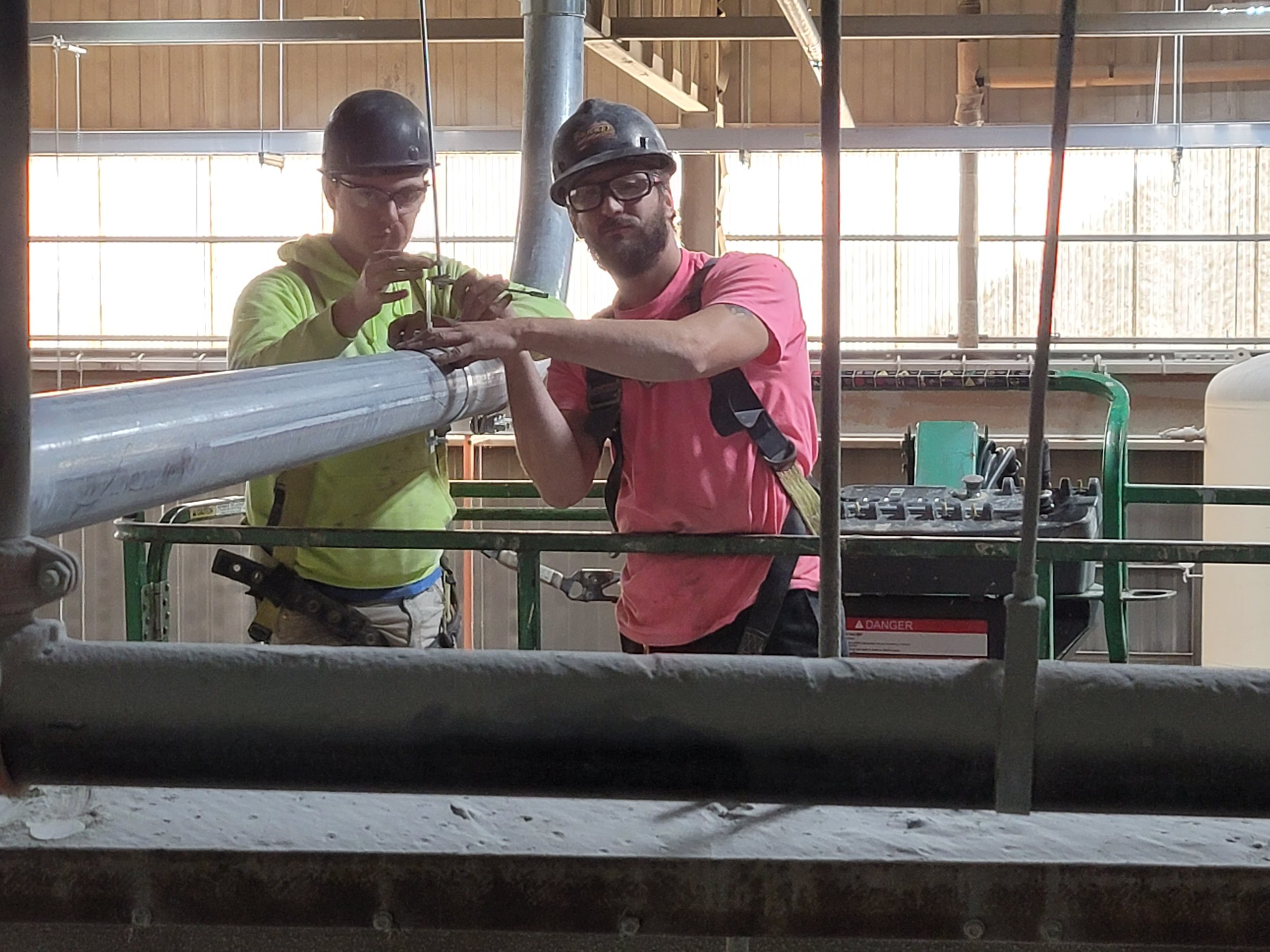 Two workers wearing helmets and safety glasses working on a construction or industrial site, handling a pipe and tools.