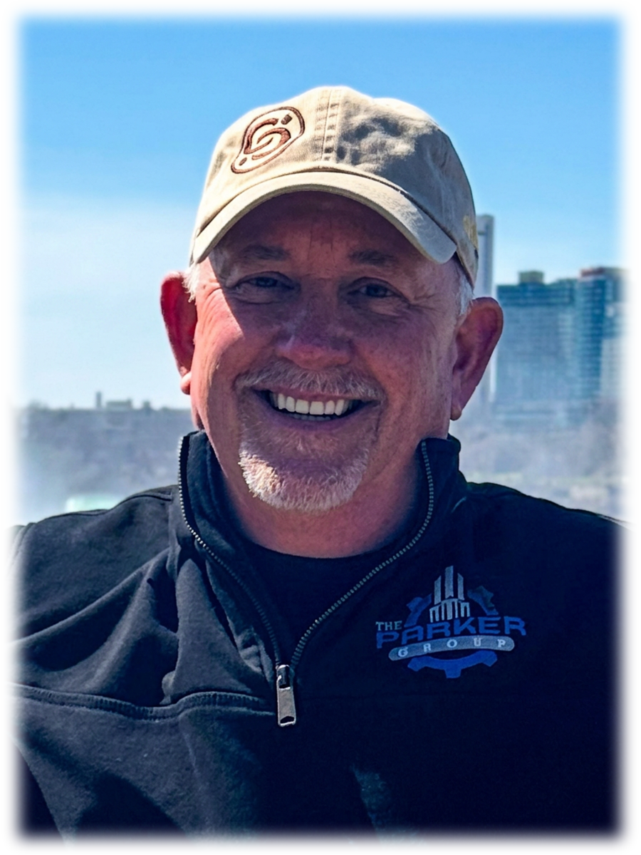 A smiling man wearing a beige cap and a black jacket with a logo that says 'The Parker Group.' He is outdoors with a blue sky and city buildings in the background.