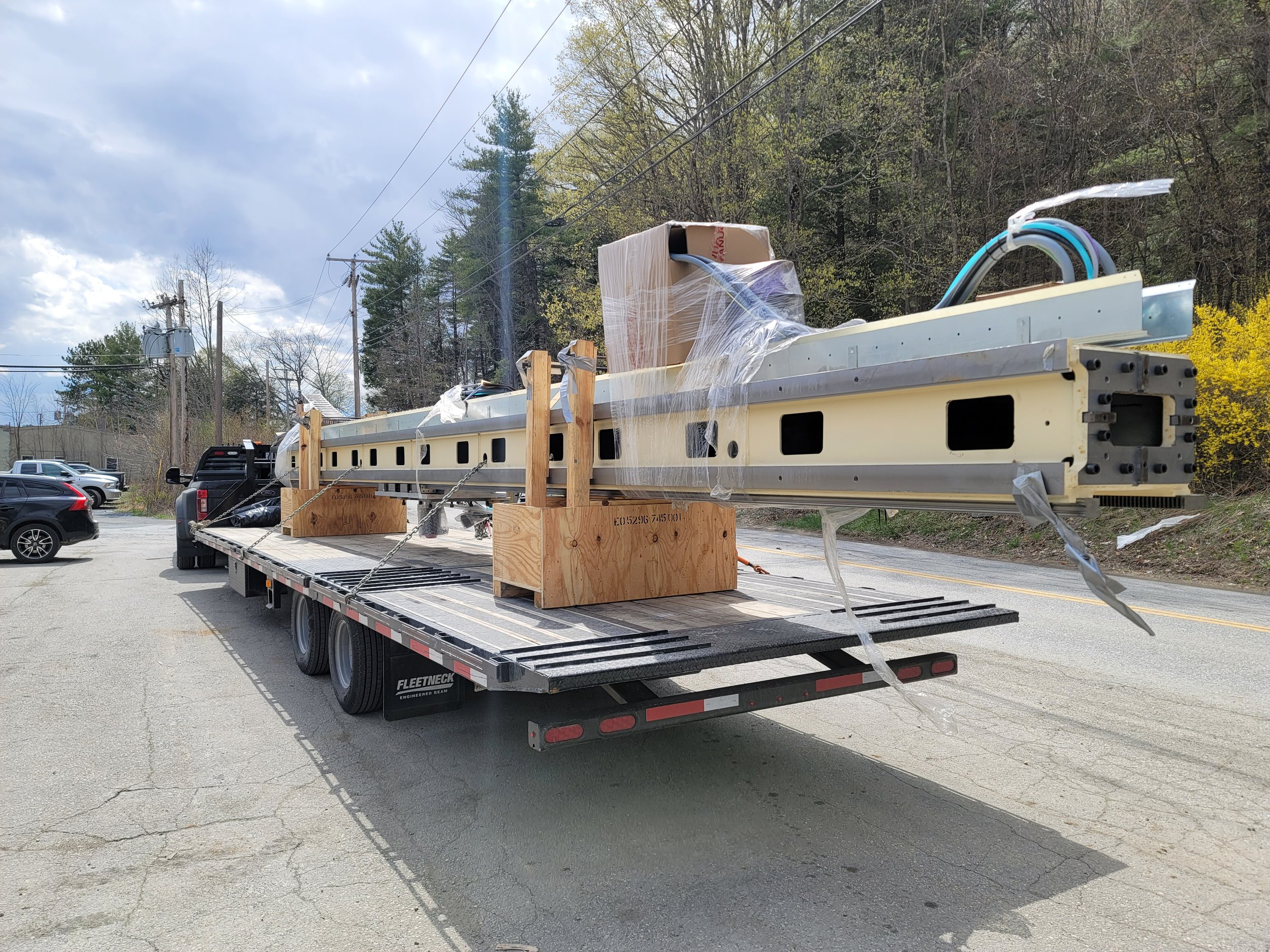 A flatbed trailer carrying large metal beams, a boxed item, and wrapped materials, parked outdoors near trees and utility poles, with parked cars and a cloudy sky in the background.