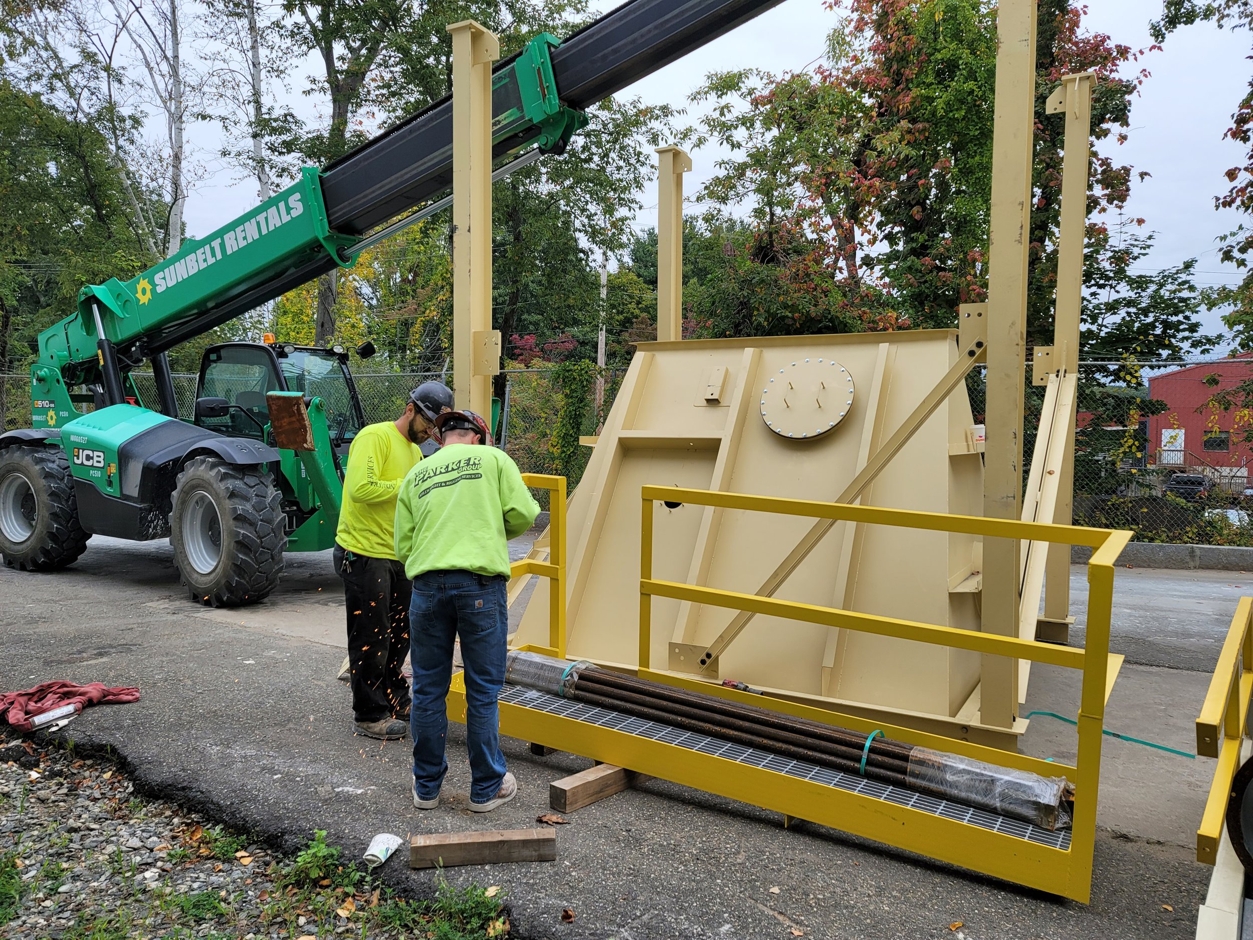 Workers in safety gear standing near yellow industrial equipment outdoors, with trees in the background.