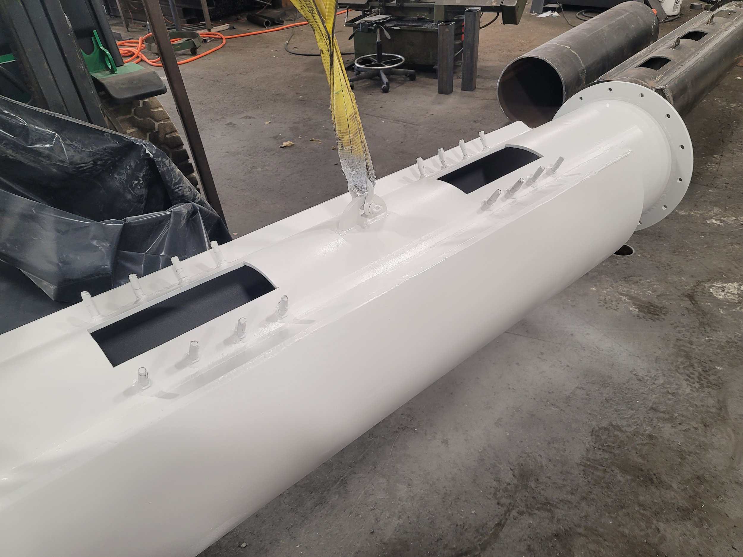 Two large hollow metal cylinders, one painted white and one unpainted, are lying on a concrete workshop floor. Various tools and equipment are scattered around, including an office chair, an orange extension cord, a workbench, and a yellow lifting st