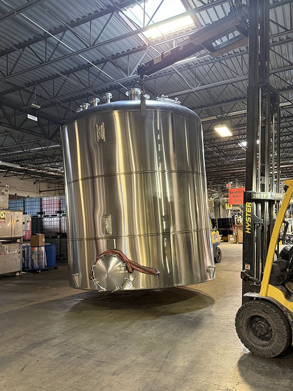 Large stainless steel tank suspended in a warehouse with forklift nearby, surrounded by industrial equipment and storage containers.