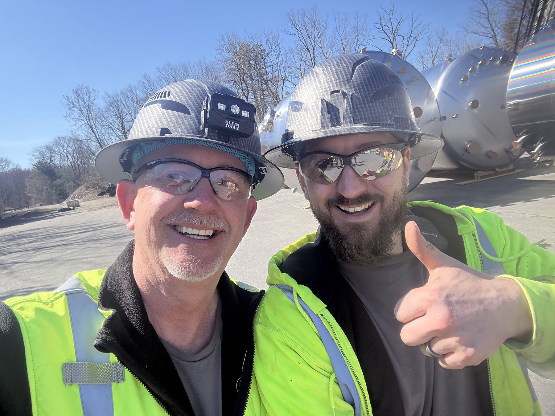 Two men wearing safety helmets, safety glasses, and high-visibility jackets taking a selfie outdoors in front of a large metallic industrial machine or equipment, with leafless trees and a clear blue sky in the background.