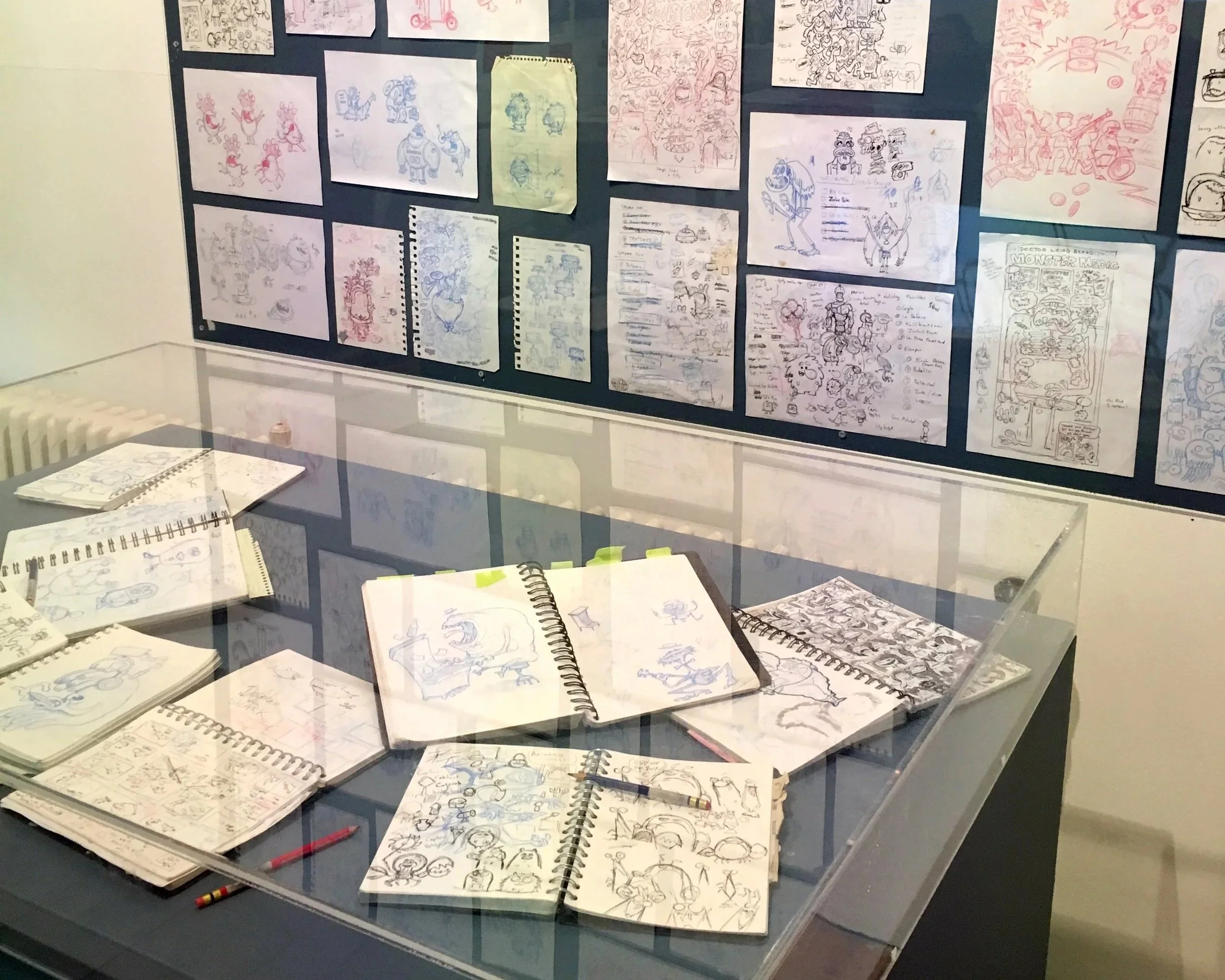 Display case with sketchbooks and loose papers containing character and scene sketches, mainly in pencil with some colored ink, organized on a black backing.