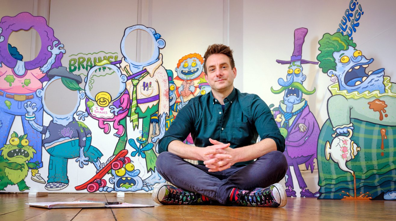 A man sitting cross-legged on the floor with colorful cartoon-style artwork of monsters and strange characters behind him.