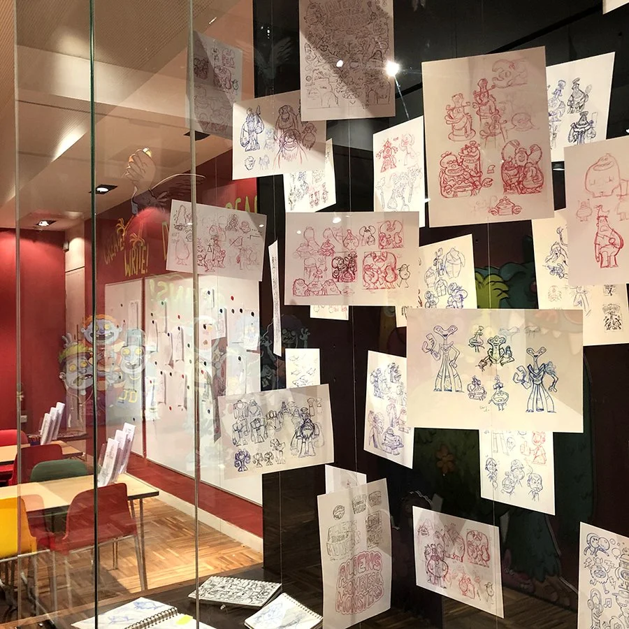 Display of various character sketches and drawings on paper hanging inside a glass enclosure, with a colorful cafe or restaurant visible in the background.