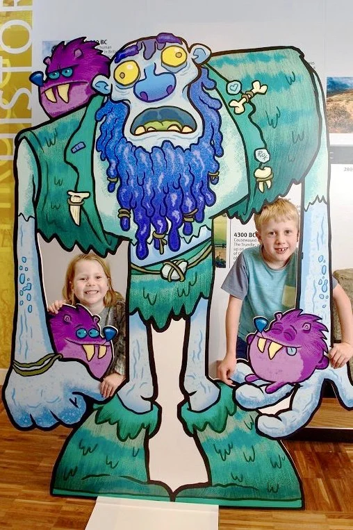 Two children stand behind a large cartoon cutout of a mystical creature with a blue beard, purple and blue face, and green robe. The cutout has openings for the children's faces, and features purple and pink monsters on its hands and shoulders.