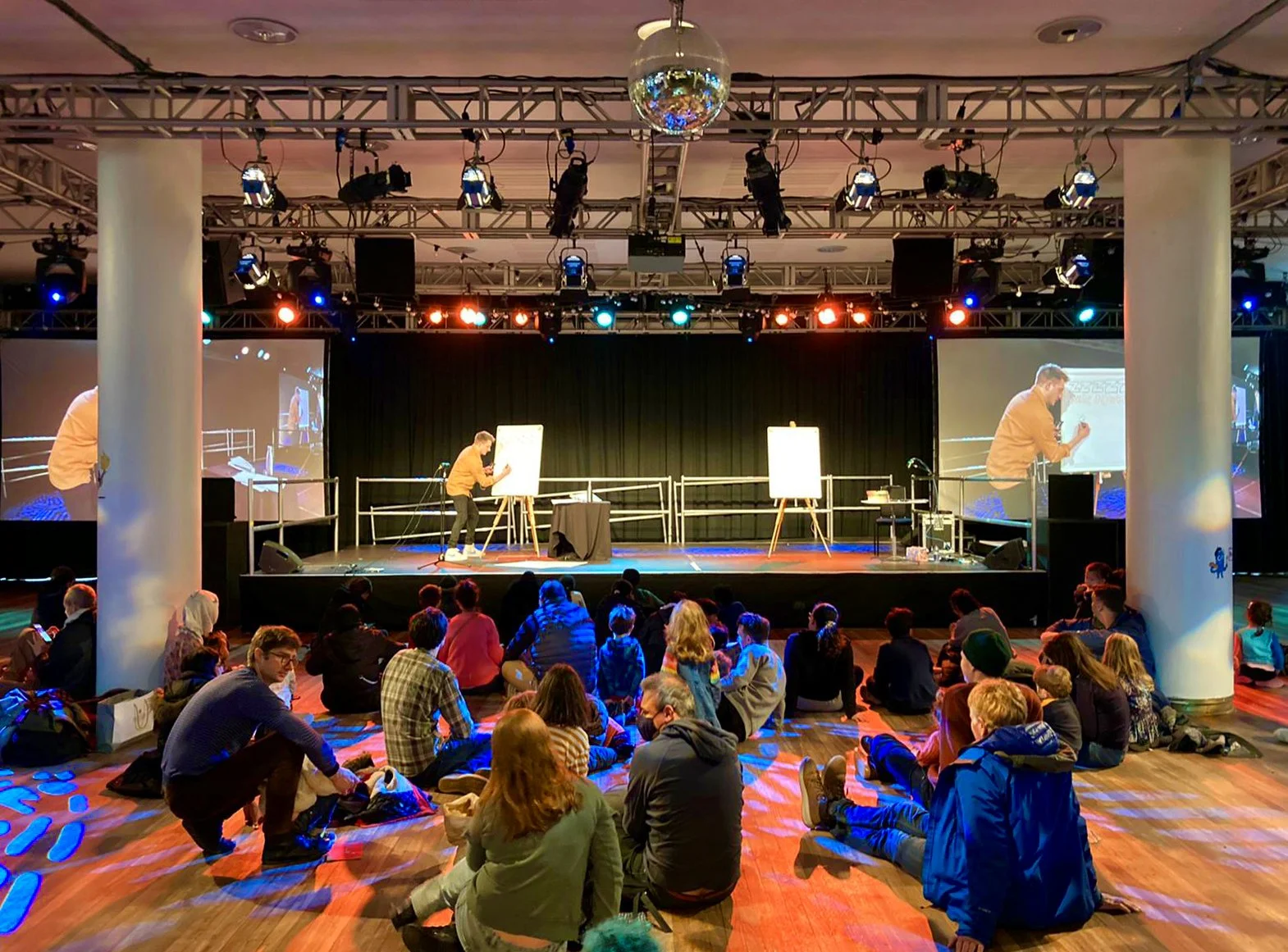 People sitting on the floor watching a presentation on stage with a person drawing on a whiteboard. Large screens on either side of the stage display the presenter. Overhead lighting and a disco ball are visible.