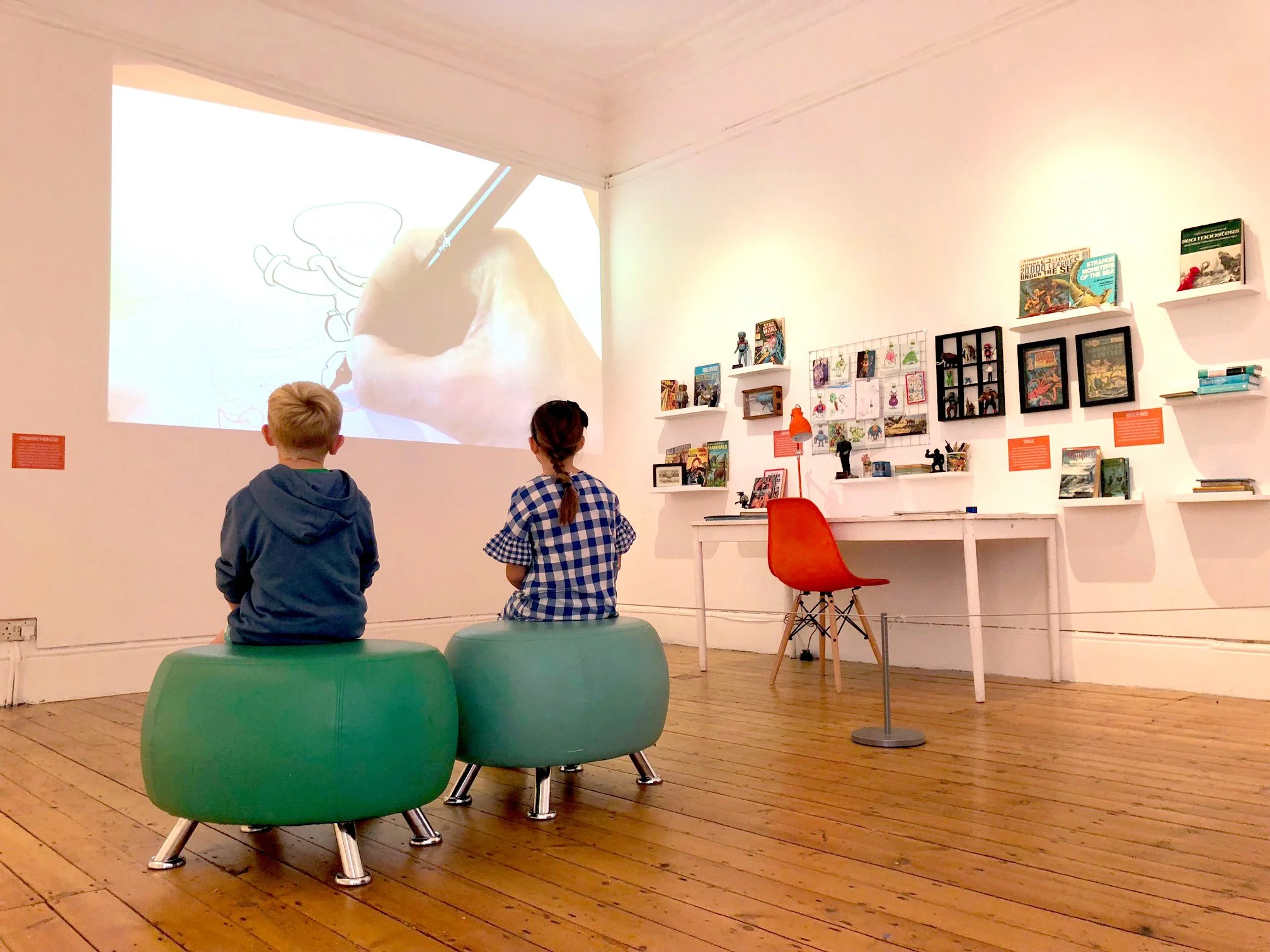 Children watching a digital drawing being projected on a white wall in an art gallery. The drawing shows a character with an oversized hand holding a stick.
