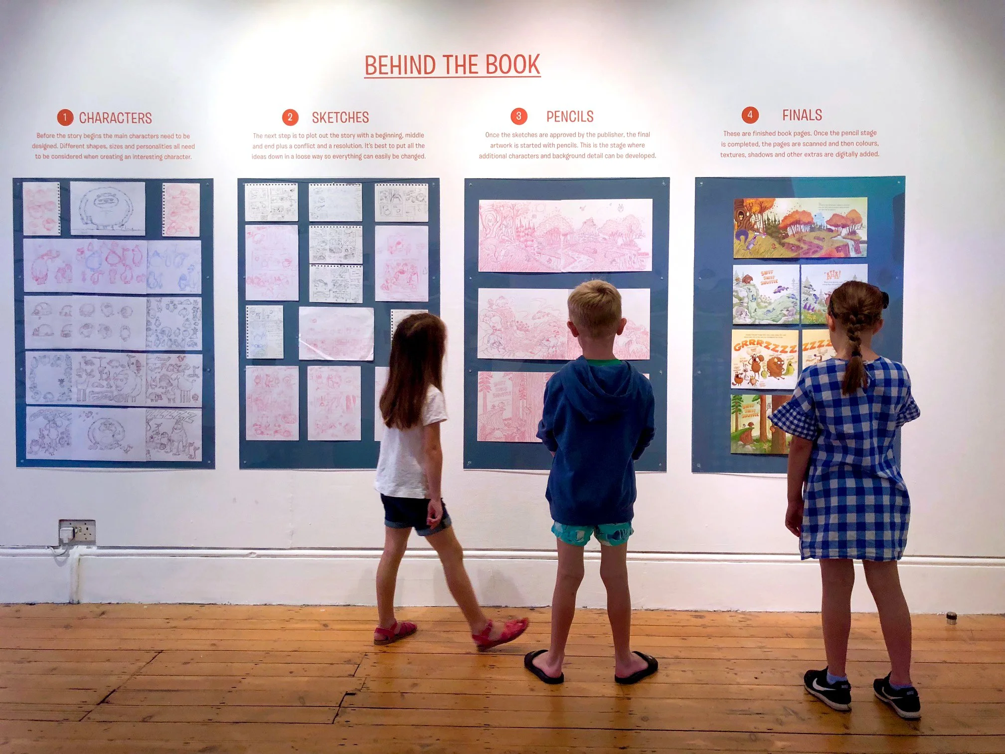 Three children standing in front of a large display wall with comic strip art and illustrations, in an indoor space with wooden floorboards.