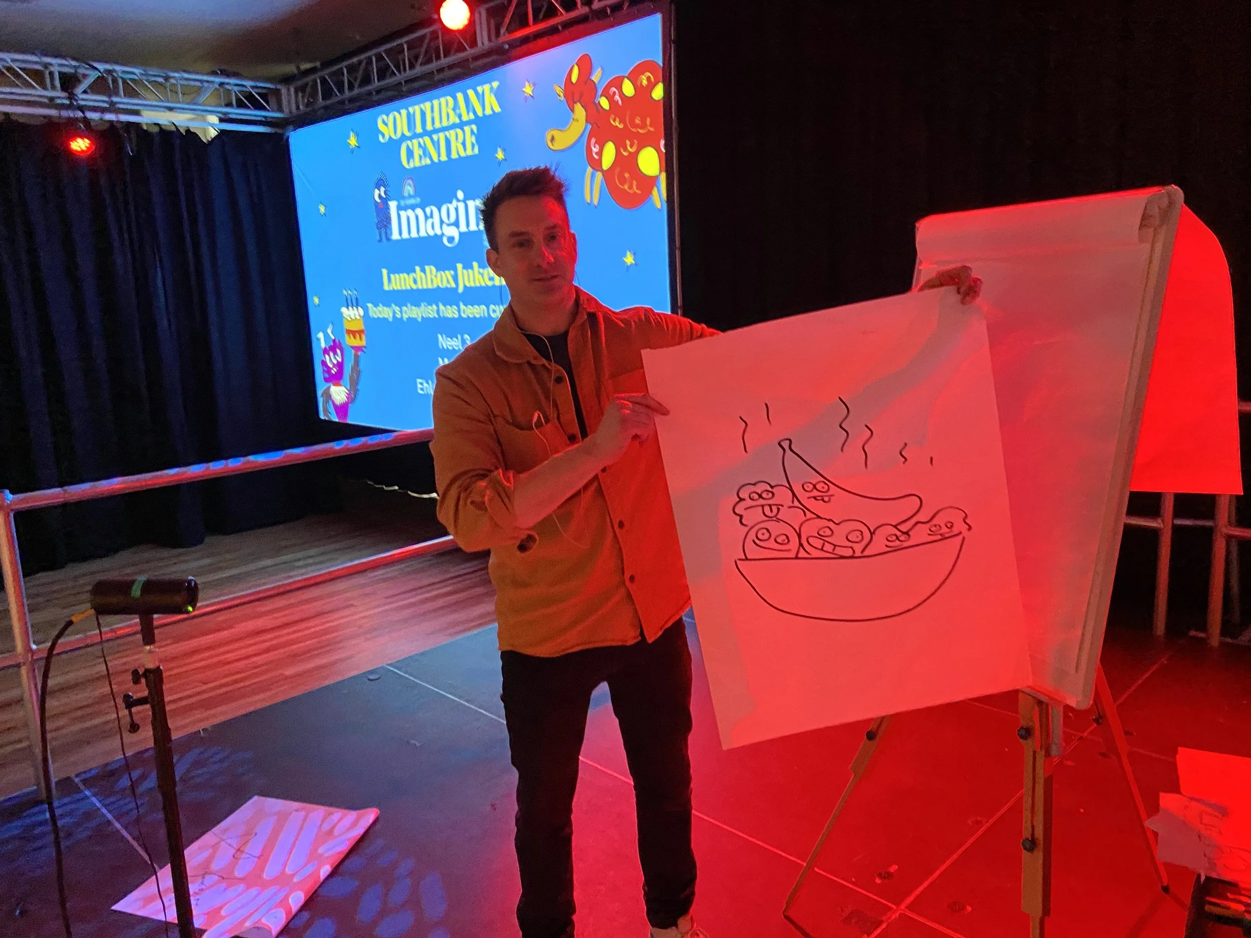 A man in a brown jacket standing next to a large orange chest with a drawing of a steaming bowl of food. He is inside a room with a stage, screen, and stage lighting, suggesting an event or presentation is taking place.