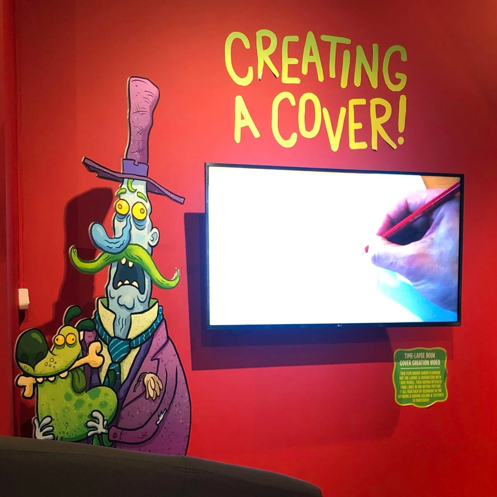 A colorful wall with cartoon characters and a TV screen. The wall has the text 'Creating A Cover!' in large, yellow, and green letters. The characters include a green creature with multiple eyes and a purple wizard with a hat, holding a smaller creature.