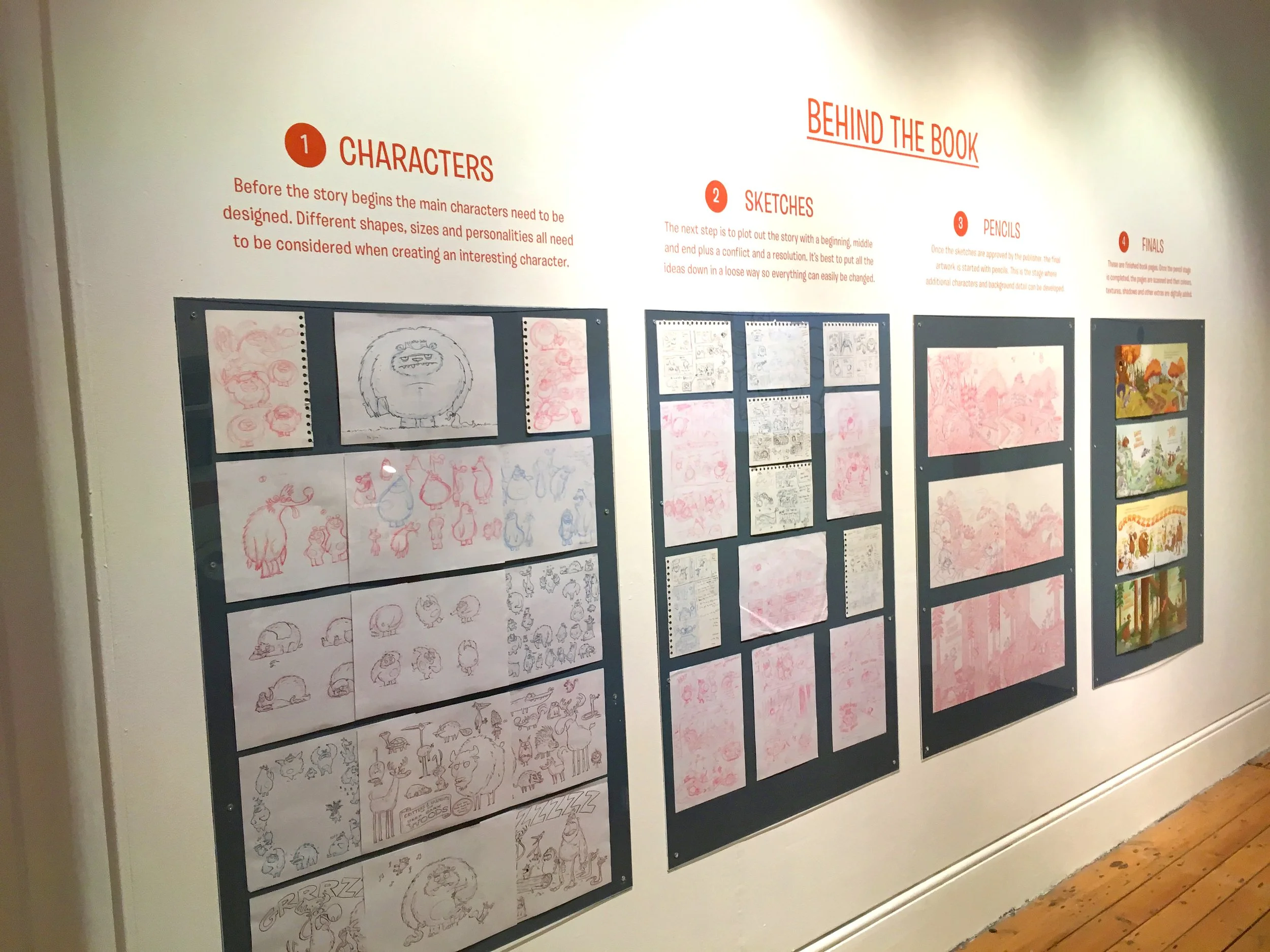 An informational wall display about the process of creating a story, including sections titled Characters, Behind the Book, Sketches, Pencils, and Finals. The display features various sketches, storyboards, and artwork.